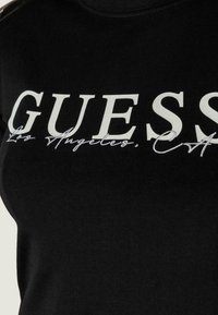 Zwarte katoenen t-shirt met een bold witte "GUESS" logo en cursieve tekst "Los Angeles, CA" eronder. Klassieke ronde halslijn en zachte textuur.
