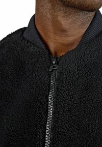 Giacca bomber in pile nera con superficie texturizzata, dotata di un colletto a costine nero e una zip nera con un tirazip con logo.