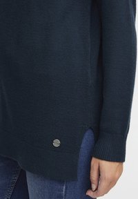 Ciemnogranatowy sweter z dzianiny z prążkowanym dołem, bocznymi rozcięciami i srebrzystym logo. Miękka tekstura i luźny krój. Nosi się go na dżinsy.