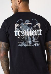 Camiseta de algodón negra con un estampado gráfico en la parte de atrás que presenta alas y la palabra "resiliente" en texto blanco en negrita, junto a los logotipos de la marca.