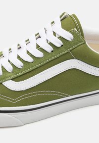 Grön canvas sneakers med vita snören och sidostrand, med sömnadsdetaljer och en svart gummisula. Texturen är jämn med en matt finish.