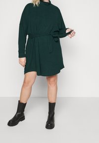Abito a maniche lunghe di colore verde scuro in morbido tessuto, con una cintura in vita. Abbinato a stivali neri fino al ginocchio, su uno sfondo neutro.