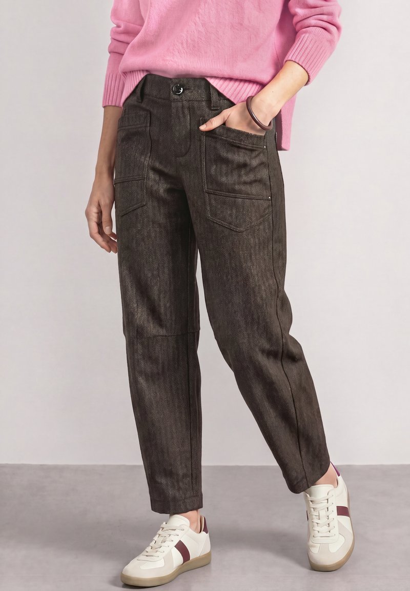 Femme portant un pantalon taille haute marron foncé avec des poches avant, un pull rose et des baskets blanches avec des rayures bordeaux.