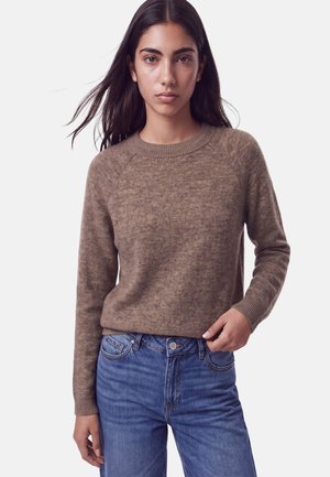 Femme aux longs cheveux foncés portant un pull en maille marron et un jean bleu, debout devant un fond clair uni, face à l'avant.