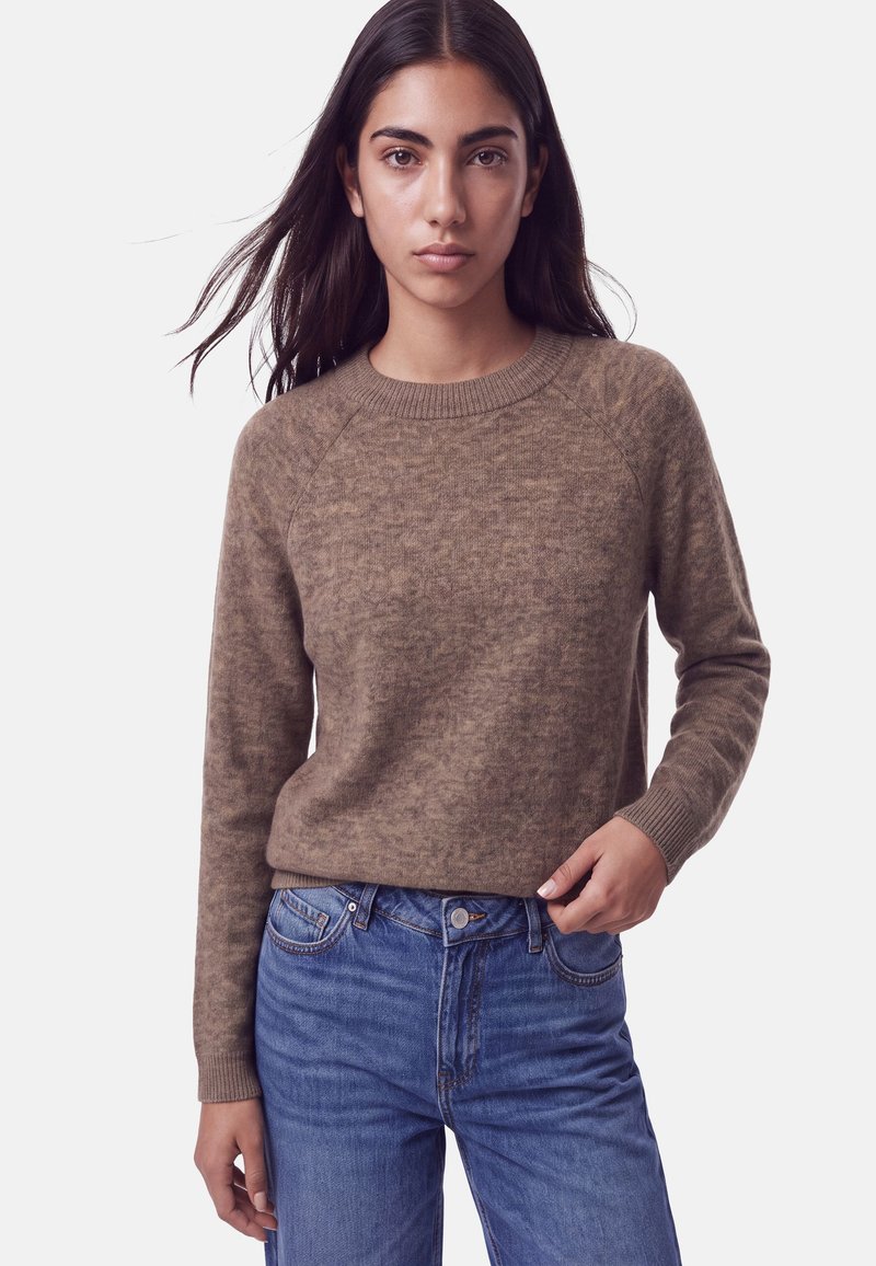 Femme aux longs cheveux foncés portant un pull en maille marron et un jean bleu, debout devant un fond clair uni, face à l'avant.
