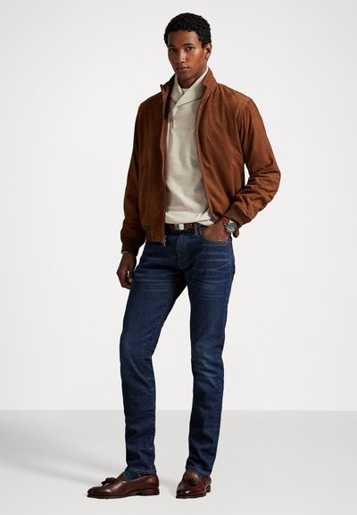 Brown suede bomber jacket, beige polo shirt, dark blue jeans, and brown loafers. Model stands with hands in pockets, showcasing outfit.