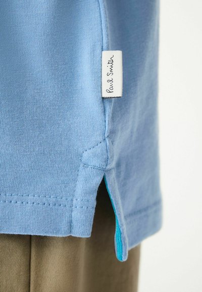 Ljusblå bomull T-shirt med sidoflikar, med en vit etikett som säger "Paul Smith." En kontrasterande turkos accent syns inuti flikarna.