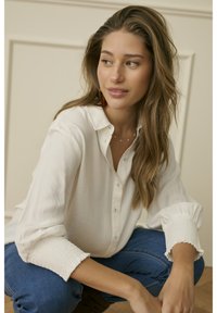 Tânără cu păr lung, îmbrăcată într-o bluză albă și blugi albaștri, stând pe podea și uitându-se spre dreapta ei.