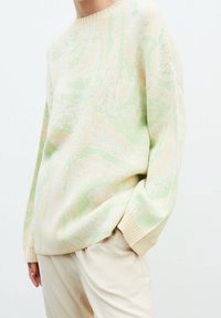 Maglione lavorato a maglia con un motivo astratto verde pastello e beige, scollatura rotonda e polsini a coste. Mostrato con pantaloni color crema.