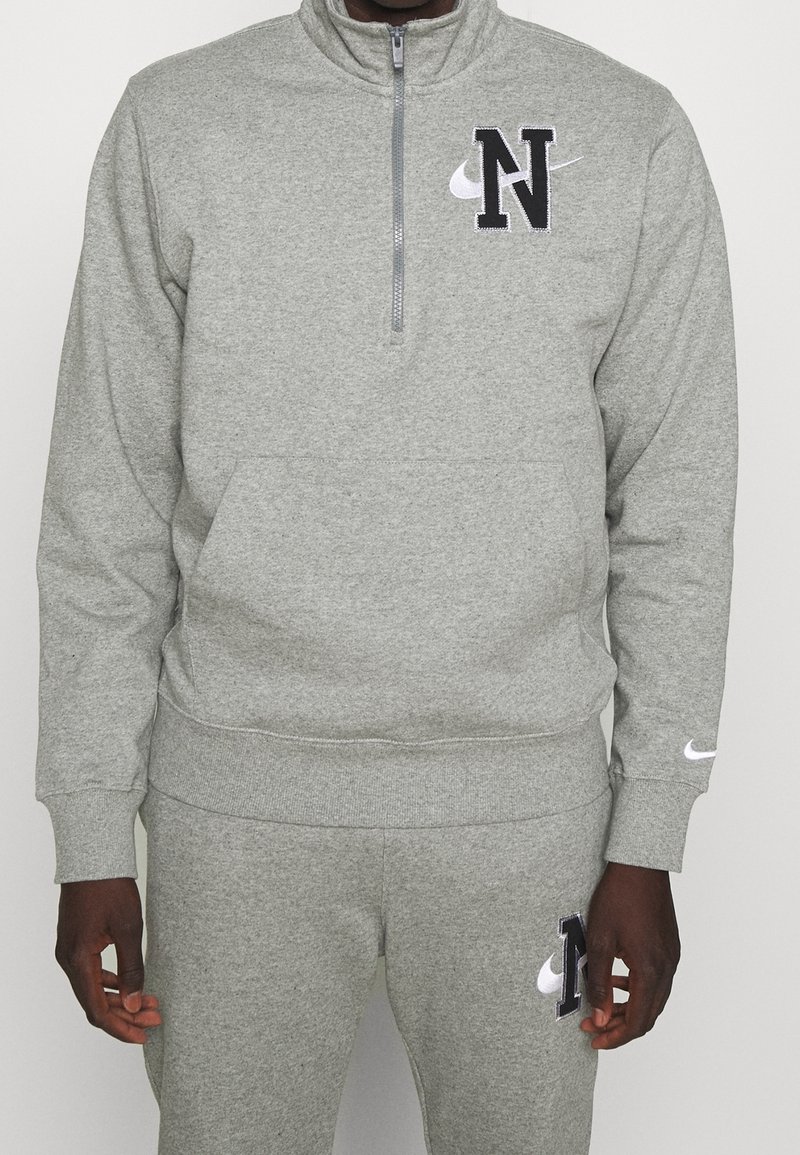 Personne portant un survêtement gris Nike avec un haut à fermeture éclair quart et un pantalon assorti, tous deux arborant une grande lettre N noire et le logo blanc Nike swoosh.