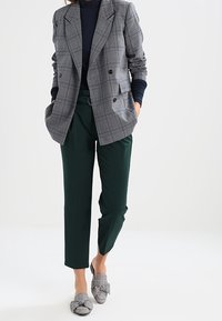 Harmaa ruudullinen kaksirivinen bleiseri, laivastonsininen pooloneule, vihreät räätälöidyt housut ja harmaat kuvioidut slip-on-kengät, joissa on rusetti.