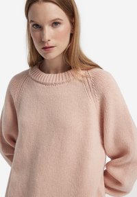 Ung kvinna med ljus hy och rakt blont hår som bär en ljusrosa stickad tröja mot en vit bakgrund.