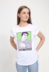 Bílý tričko s grafickým designem svalnaté postavy, na zeleném pozadí a textem „THE HULK“ v tučných černých písmenech. Volný střih.