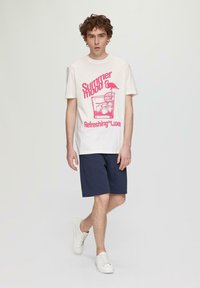 Camiseta de algodón con fondo crema, que presenta un estampado gráfico en rosa de una bebida con un flamenco. Combinada con pantalones cortos navy y zapatillas blancas.