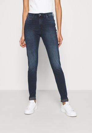Femme portant un jean slim bleu foncé avec des revers et des baskets blanches, debout devant un fond clair uni.