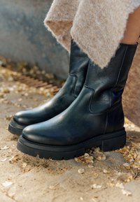 Botas de tobillo de cuero negro con un acabado suave, suela de goma gruesa y detalle en el panel lateral. Colocadas sobre un fondo arenoso y texturizado con hojas.