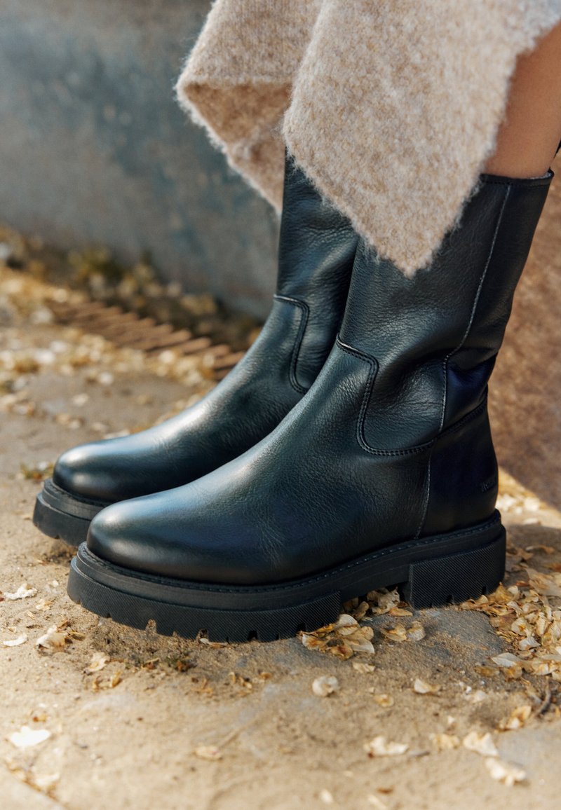 Botas de tobillo de cuero negro con un acabado suave, suela de goma gruesa y detalle en el panel lateral. Colocadas sobre un fondo arenoso y texturizado con hojas.