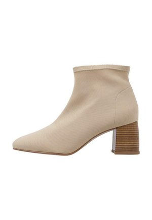 Botín beige hasta el tobillo con una parte superior de tela elástica, puntera afilada y un tacón de madera a rayas. Textura suave y diseño minimalista.