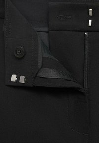 Gros plan d’un pantalon noir sur mesure montrant une fermeture éclair partiellement ouverte, un bouton noir et deux agrafes argentées.