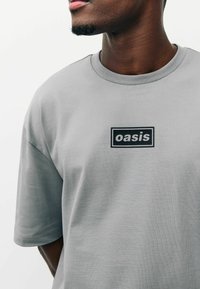 Szürke rövid ujjú póló, puha pamutból készült, közepén fekete, "oasis" felirattal ellátott téglalap alakú logóval.