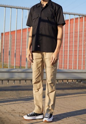 Personne portant une chemise noire à manches courtes avec boutons, un pantalon cargo beige et des baskets noir et blanc, debout sur un sol pavé près d'une clôture métallique.