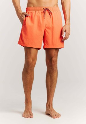 Homme debout pieds nus portant un short de bain orange vif avec poches latérales et taille à cordon.
