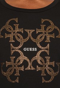 Mustassa t-paidassa on silmiinpistävä kultakuvioitu logomuotoilu, jossa on toisiinsa kytkeytyviä muotoja ja sana "GUESS" valkoisena keskellä.