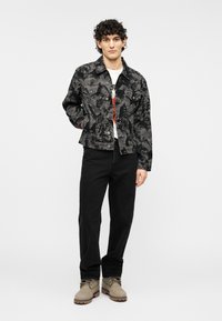 Μαύρο τζιν μπουφάν με διακριτικό υφασμάτινο μοτίβο, φορεμένο πάνω από λευκό μπλουζάκι με γραφικά, συνδυασμένο με μαύρο παντελόνι και μπεζ μπότες.