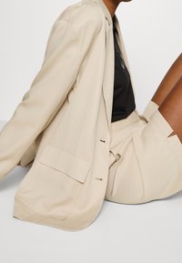 Beige blazer med en afslappet pasform, der har en enkelt lomme, minimalt hardware, glat tekstur, og som er parret med en sort top og en beige nederdel.