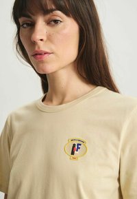 T-shirt en coton beige avec un col rond, des manches courtes et un patch logo coloré sur la poitrine comprenant un graphique et du texte.