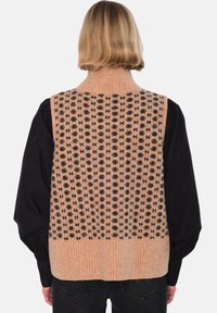 Strikket vest i ferskenfarge med svart geometrisk mønster, med ribbet krage og kant. Stoffets tekstur virker myk og koselig.