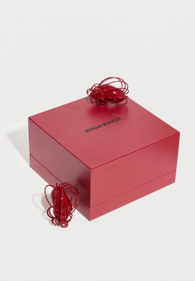 Rdeča škatla Hugo Kreit z dvema rdečima uhanoma iz žice v obliki okraskov, eden na vrhu, drugi ob strani škatle, na belem ozadju.