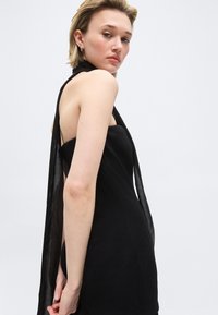 Ung kvinde med kort blondt hår iført en ærmeløs sort kjole med et gennemsigtigt tørklæde draperet over skulderen, der kigger over skulderen.