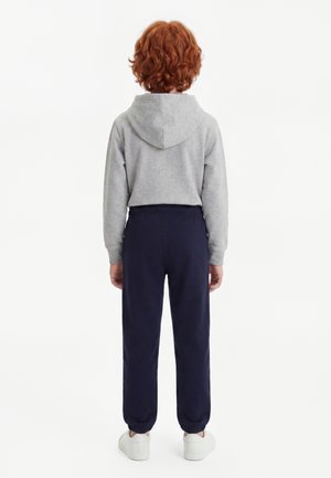 Kind met rood krullend haar staat met de rug naar ons toe, draagt een grijze hoodie, donkerblauwe joggingbroek en witte sneakers op een witte achtergrond.