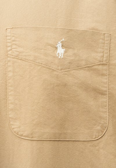 Smėlio spalvos audinio kišenė su detaliu siuvimu ir mažu baltu užsiuviniu, vaizduojančiu polo žaidėją, laikantį lazdą, centrinėje vietoje virš kišenės angos.