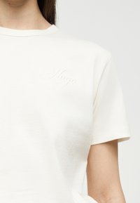 Γυναίκα που φοράει απλό λευκό κοντομάνικο μπλουζάκι με διακριτικό ανάγλυφο κείμενο "Hugo" στο πάνω αριστερό μέρος του στήθους.