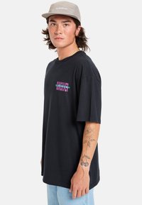 Camiseta de algodón negra con mangas cortas y escote tipo crew, que presenta un logo colorido en la parte delantera. El modelo lleva unos jeans de color azul claro.