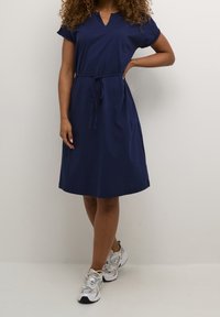 Tmavě modré bavlněné šaty s krátkými rukávy, výstřihem do V, nastavitelným opaskem a áčkovým střihem, doplněné bílými a šedými sportovními botami.