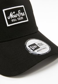 Black adjustable cap made of textured fabric featuring a white embroidered logo on a rectangular patch and a circular New Era tag.