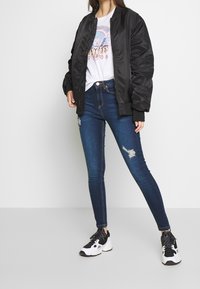 Černá bomber bunda s žebrovanými manžetami, tričko s grafickým potiskem, roztrhané modré skinny džíny a černobílé tenisky.
