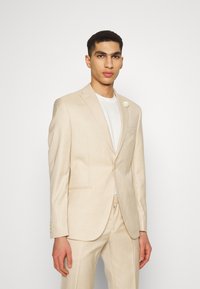 Sacou bej personalizat, cu un blazer cu un singur nasture, rever în formă de tăietură și un buzunar pentru piept. Asortat cu un tricou alb și un ac de cravată decorativ.