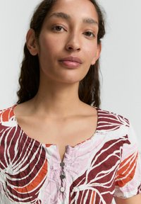 Robe à manches courtes avec un col rond, arborant un motif floral dans des teintes de rouge, rose et blanc. Détail de patte de boutonnage à trois boutons.