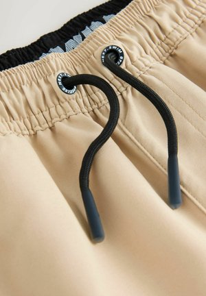 Shorts de bain beige avec une taille élastique, un cordon noir et une doublure bleue à motifs. Comprend des œillets renforcés et une texture lisse.