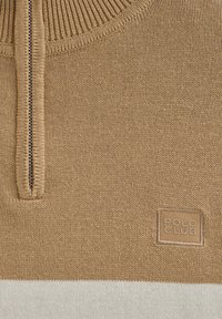 Suéter de punto beige con cuello de media cremallera, que presenta una franja blanca en contraste y un parche de logo cosido en la parte inferior izquierda.