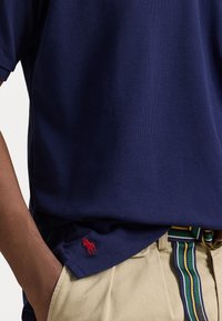 Pánské polo triko navy barvy z texturované tkaniny s krátkým rukávem, se malým vyšitým červeným logem na lemu. V kombinaci s béžovými kalhotami a pruhovaným opaskem.