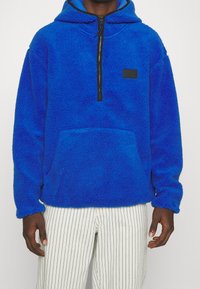 Pullover in pile blu con mezza zip e cappuccio. Presenta una tasca frontale e un'etichetta con logo nera. Texture morbida con polsini e orlo a costine.