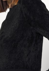 Femme portant un pull noir texturé à manches longues, cheveux bruns lisses partiellement visibles sur un fond blanc.