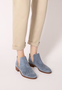 Helle blauene Wildleder-Stiefeletten mit niedrigem Absatz und seitlichen Ausschnitten werden mit beigen, umgeschlagenen Hosen kombiniert. Sanfte Textur und elegantes Design.
