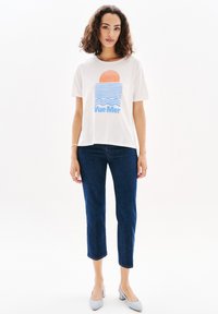 T-shirt bianco a maniche corte con un design grafico di un sole e onde in arancione e blu, abbinato a jeans cropped di denim scuri e scarpe azzurre chiare.