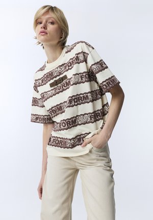 Young person with short blonde hair wears cream and brown patterned t-shirt and light beige pants, standing with one hand in pocket.